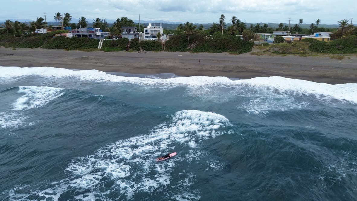 Ramon on a pink bodyboard among breaking waves, with Arenas Negra beyond the black-sand beach